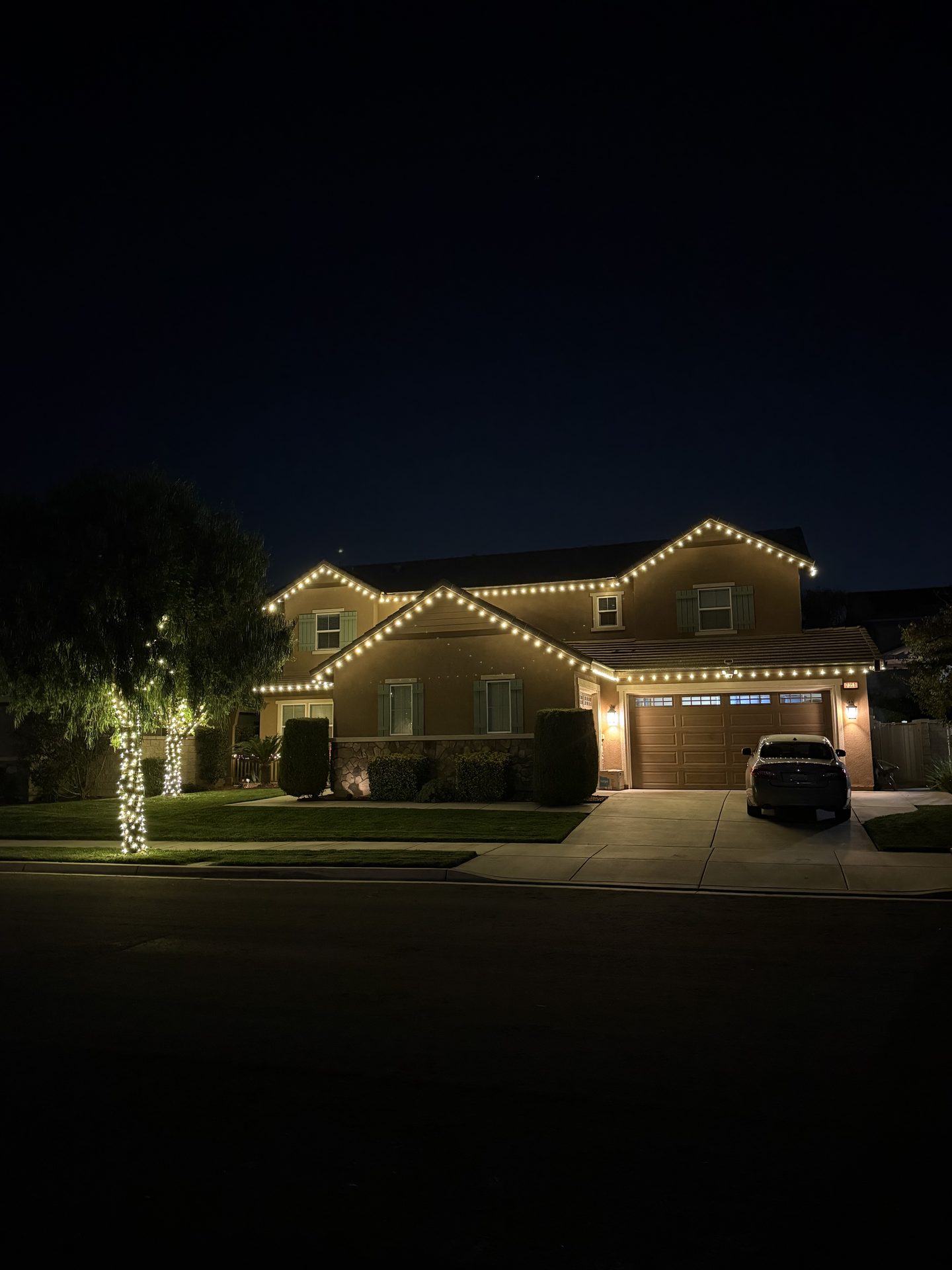 Two-story home with warm white roofline lighting and wrapped tree at night