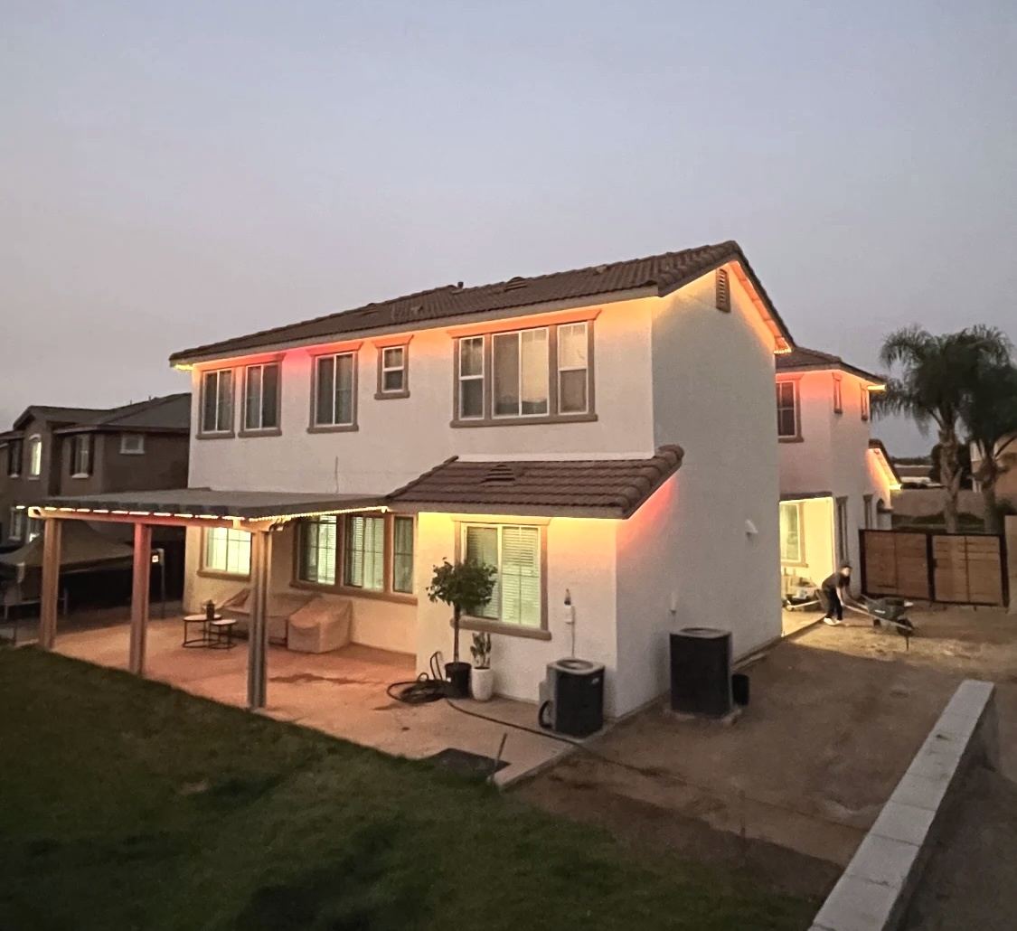 Two-story home with warm white permanent roofline lighting at sunset, backyard patio view