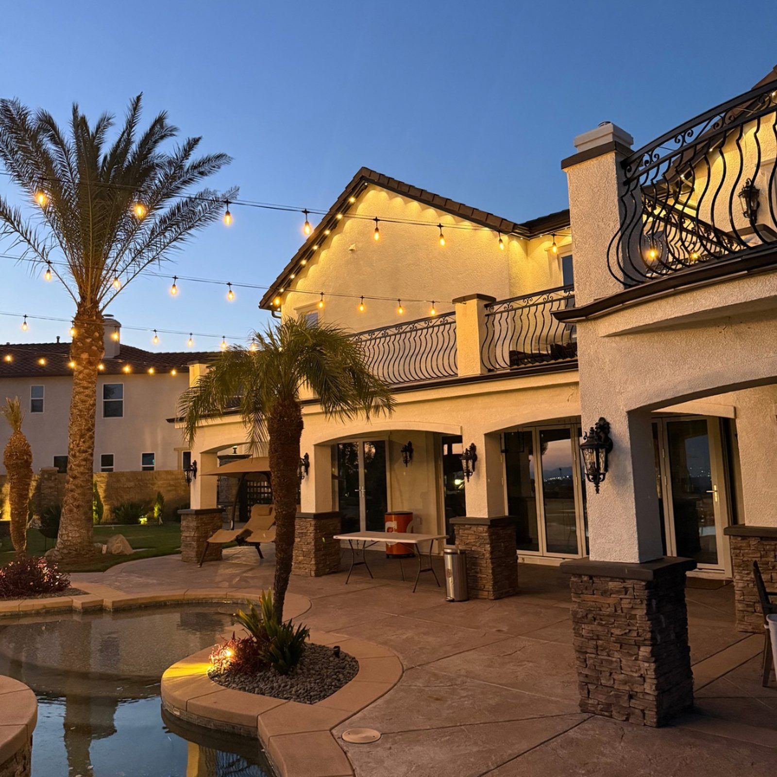 String lighting over backyard pool with palm trees and balcony at dusk