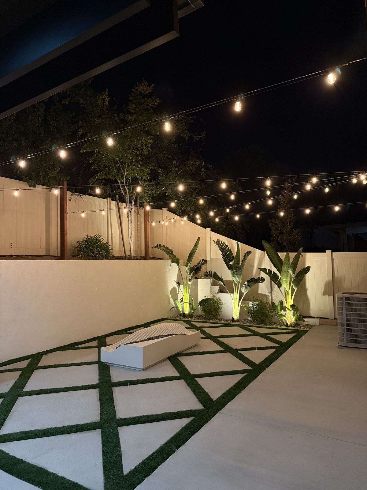 String lighting over backyard patio with turf pattern and birds of paradise at night